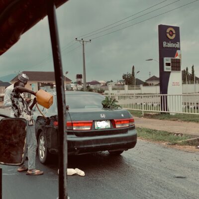 A man fills petrol from a container into a car by a Rainoil station, on a rainy street.
