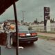 A man fills petrol from a container into a car by a Rainoil station, on a rainy street.
