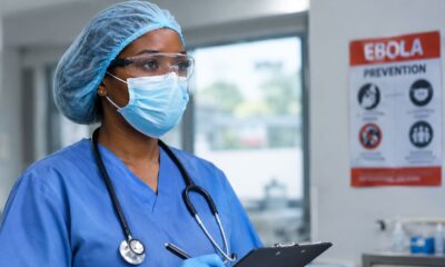 Medical worker in protective scrub cap, safety goggles, face mask, and gloves holding a clipboard near an Ebola prevention poster.