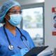 Medical worker in protective scrub cap, safety goggles, face mask, and gloves holding a clipboard near an Ebola prevention poster.