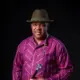 Veteran Nollywood actor Francis Duru wearing a purple striped traditional shirt, fedora hat, and holding a walking stick.