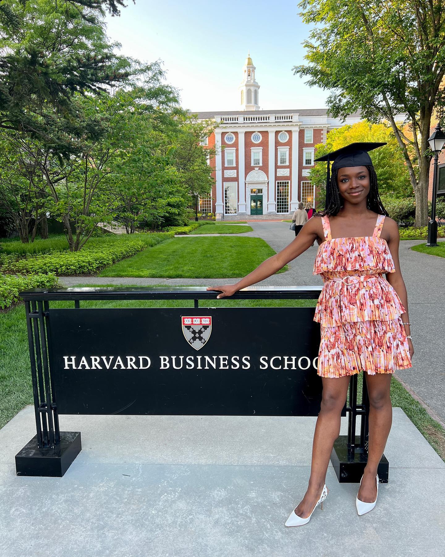 HaloBraid founder Yinka Ogunbiyi wearing a patterned dress and graduation cap beside the Harvard Business School sign.