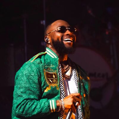 Davido holding a microphone on stage wearing a green lace varsity bomber jacket, sunglasses, and layered diamond chain necklaces during a live concert in London.