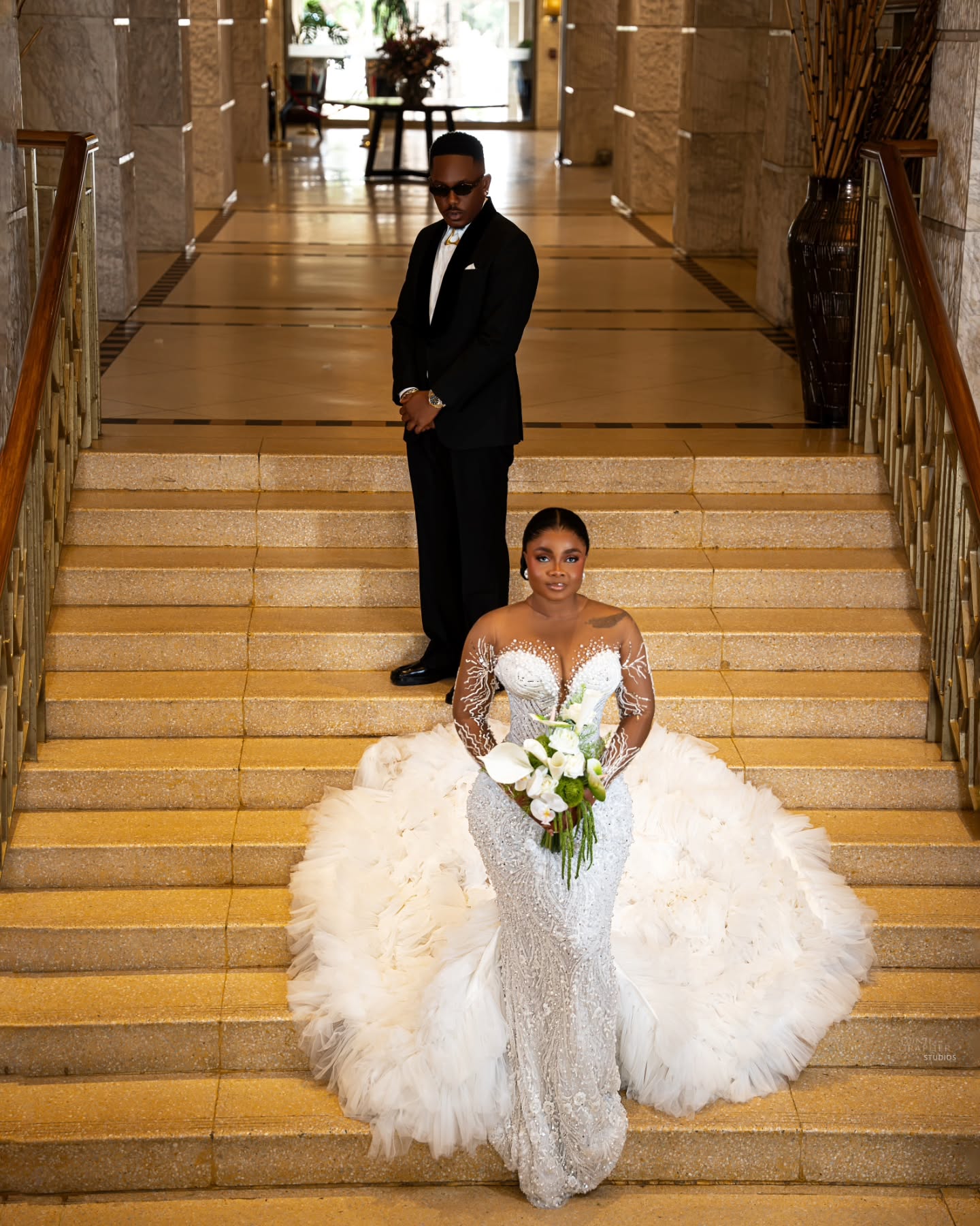 Bimbo Ademoye in a pearl embellished mermaid bridal gown with ruffled train standing on stairs in front of Timini Egbuson
