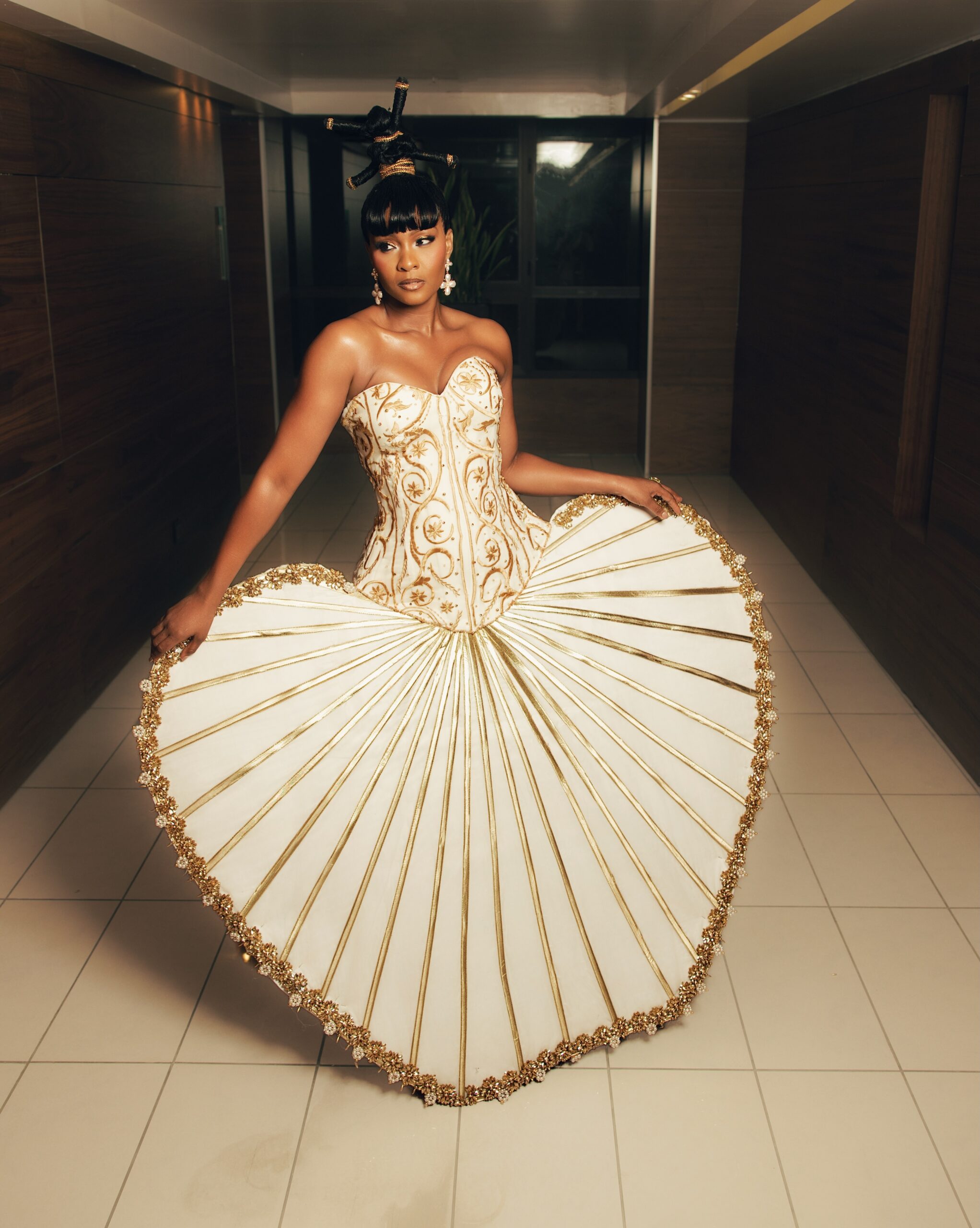 Osas Ighodaro standing in a hallway wearing an inverted heart fan-pleated skirt for BellaNaija at 20 Grand Red Carpet Experience.