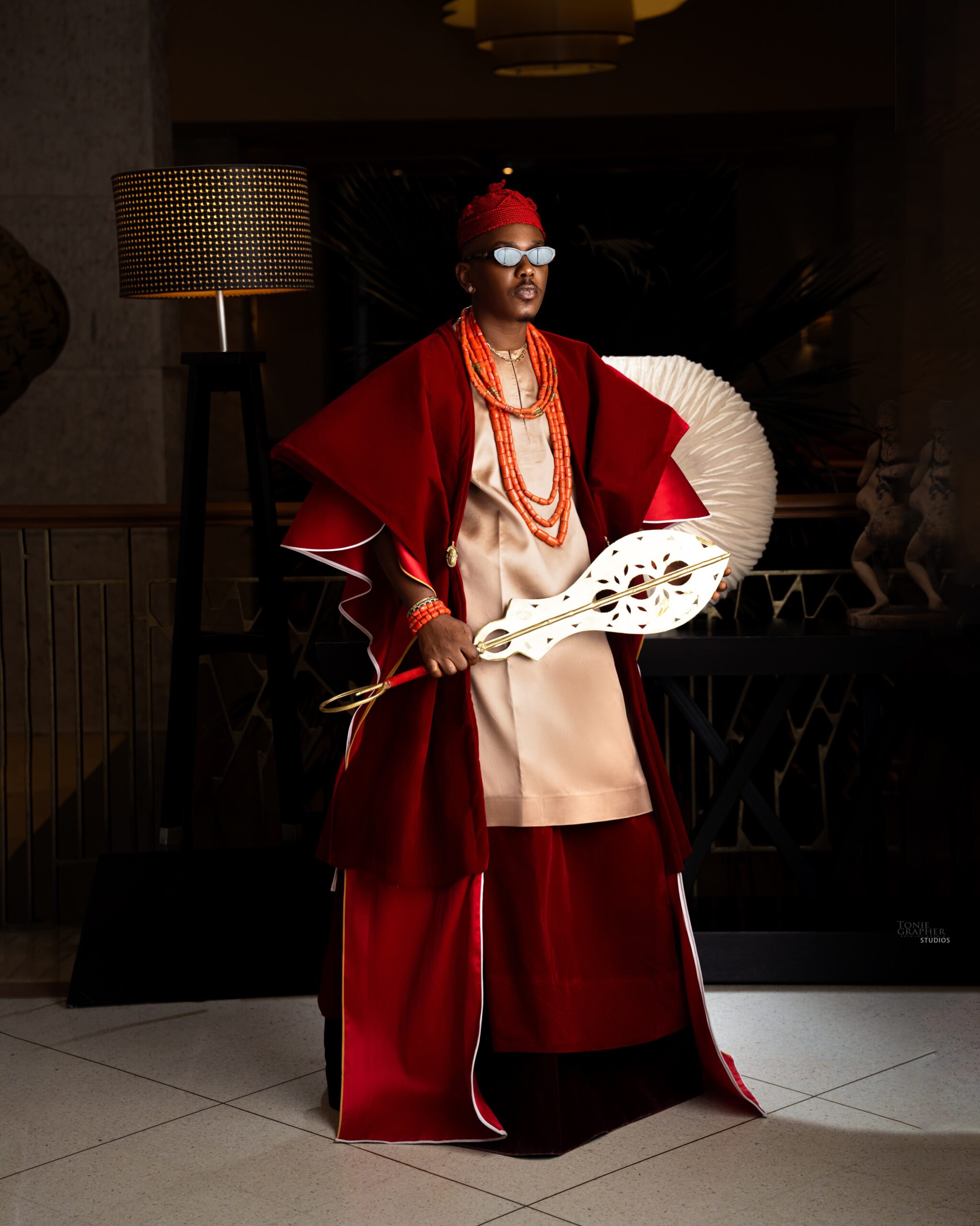 Timini Egbuson wearing a gold tunic red velvet robe coral beads sunglasses and carved Akupe hand fan