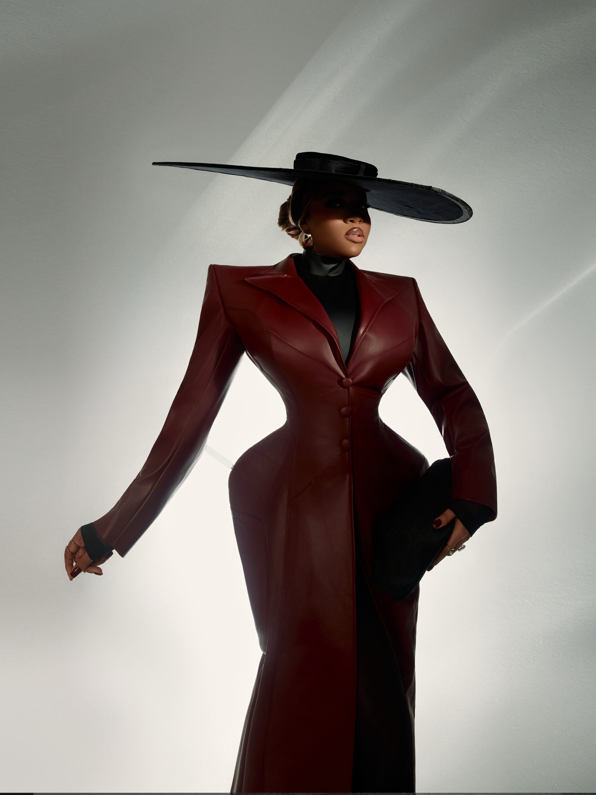 Veekee James in a sculpted oxblood leather coat-gown by Abbas Woman featuring padded shoulders, a high-neck black turtleneck, and an oversized black wide-brim hat.