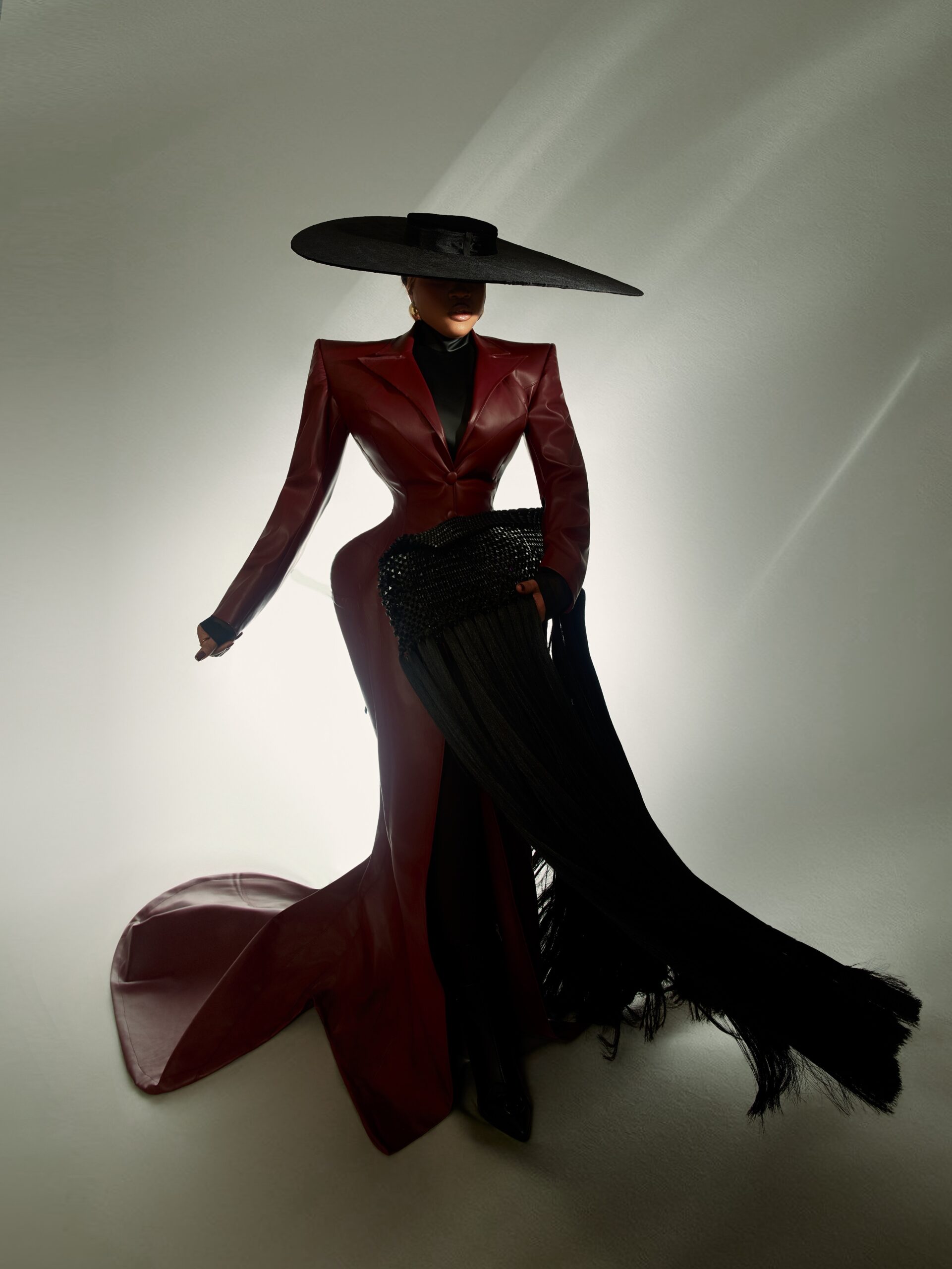 Veekee James showcasing a black beaded fringe clutch bag, custom oxblood leather coat-gown by Abbas Woman, and an oversized black hat for the BellaNaija at 20 High Fashion Icon theme.