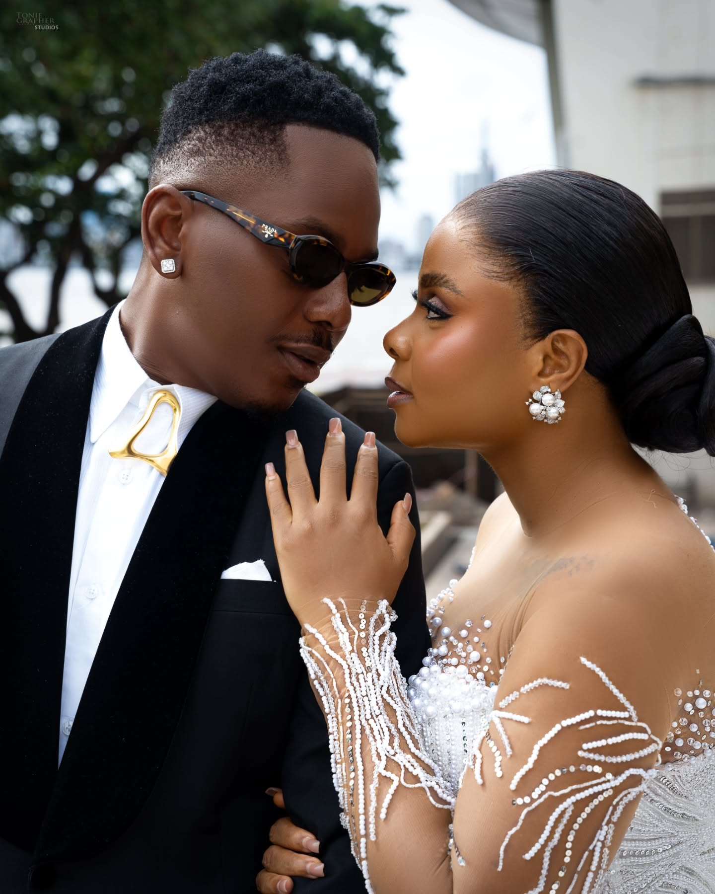 Timini Egbuson in a black tuxedo and gold collar brooch beside Bimbo Ademoye in a pearl embellished bridal gown