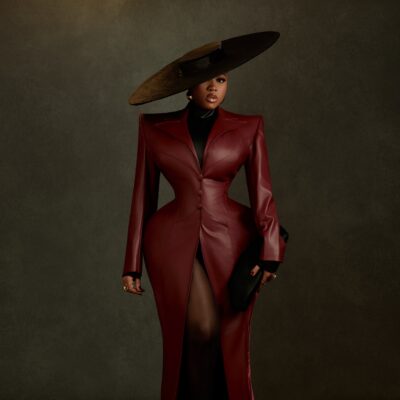 Veekee James wearing an oxblood leather coat-gown by Abbas Woman with structured shoulders, peplum hips, a wide-brim hat, and pointed heels at the BellaNaija at 20 Grand Red Carpet Experience.