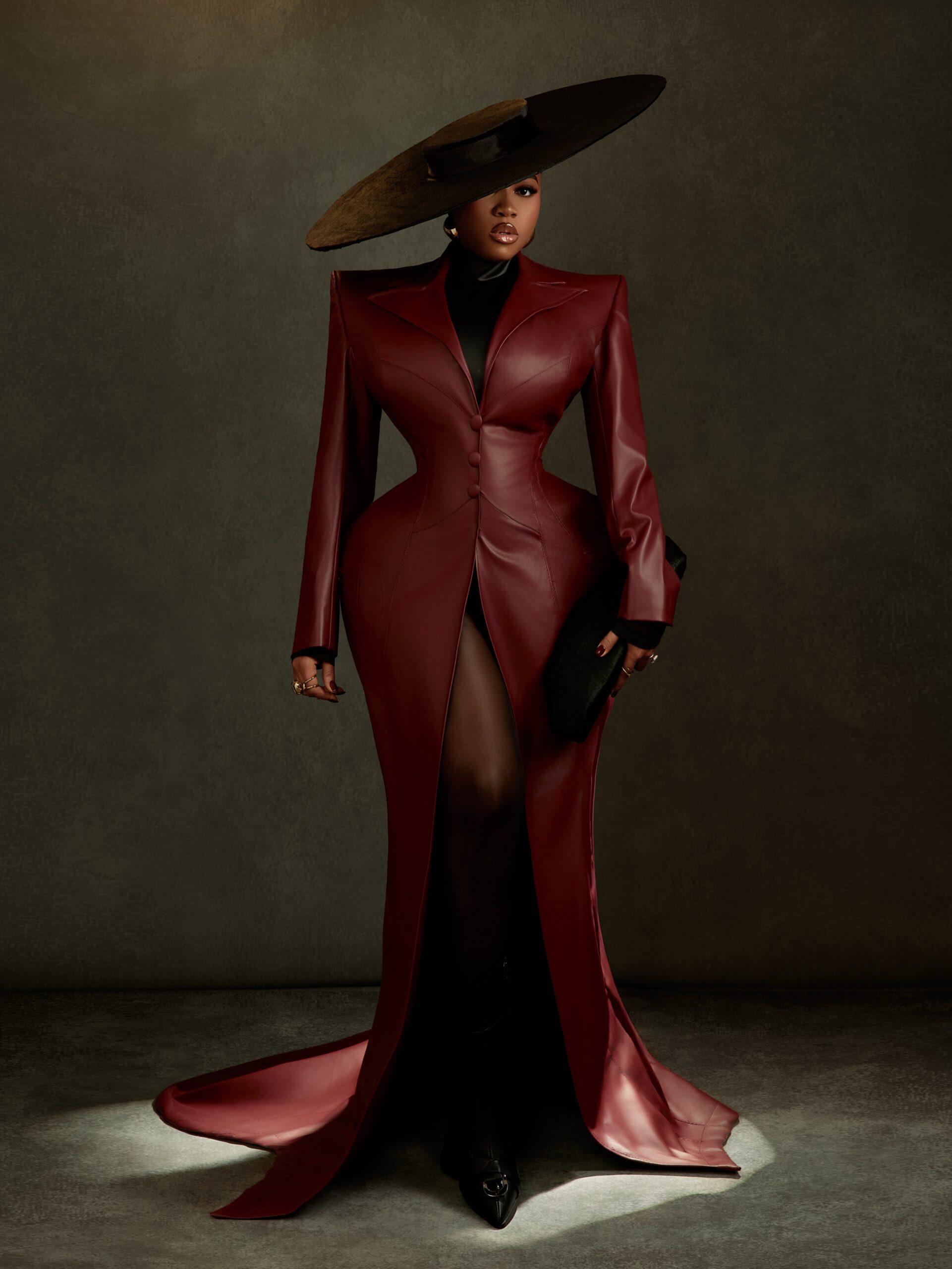 Veekee James wearing an oxblood leather coat-gown by Abbas Woman with structured shoulders, peplum hips, a wide-brim hat, and pointed heels at the BellaNaija at 20 Grand Red Carpet Experience.