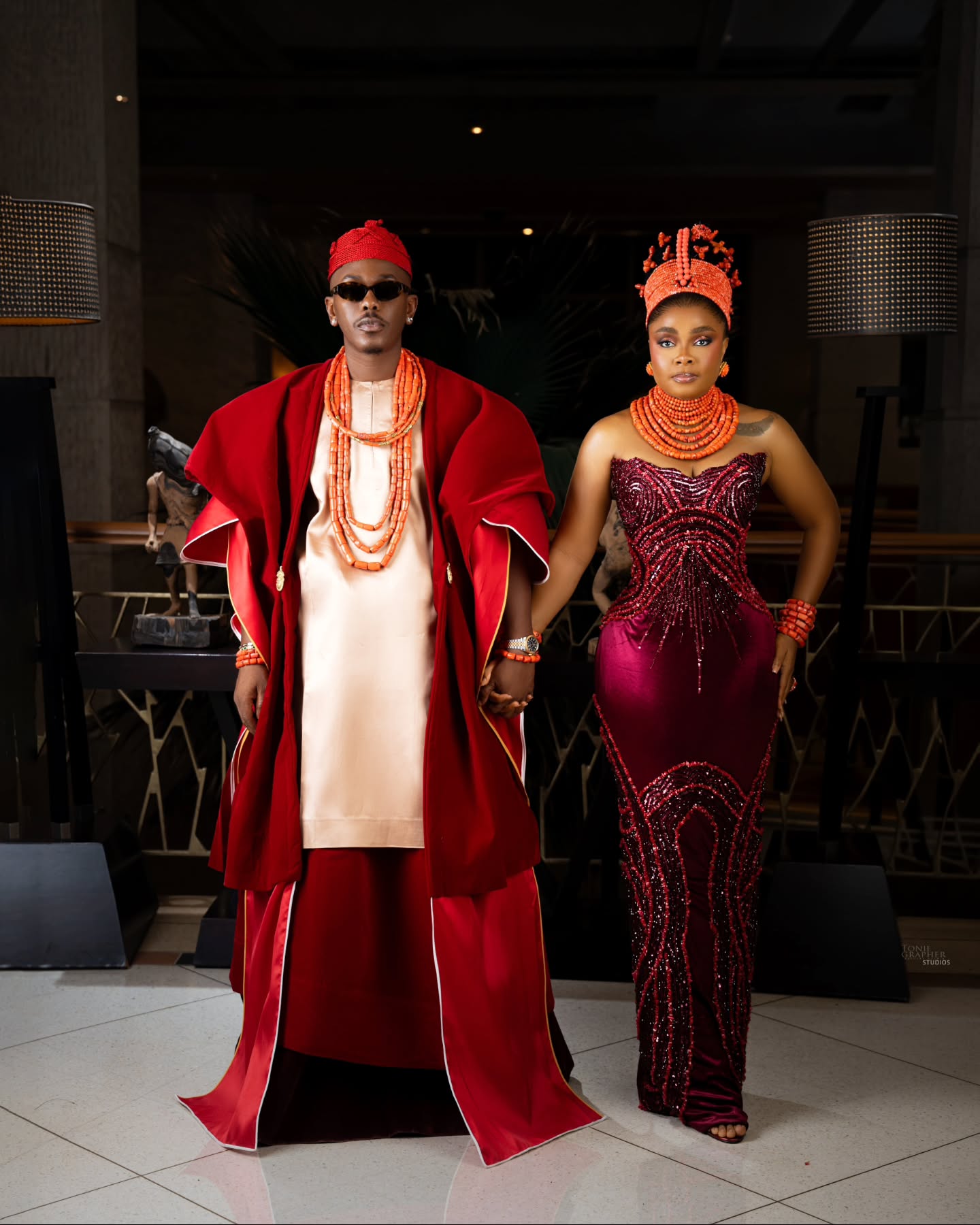 Timini Egbuson in a red velvet robe beside Bimbo Ademoye in a maroon beaded gown holding hands
