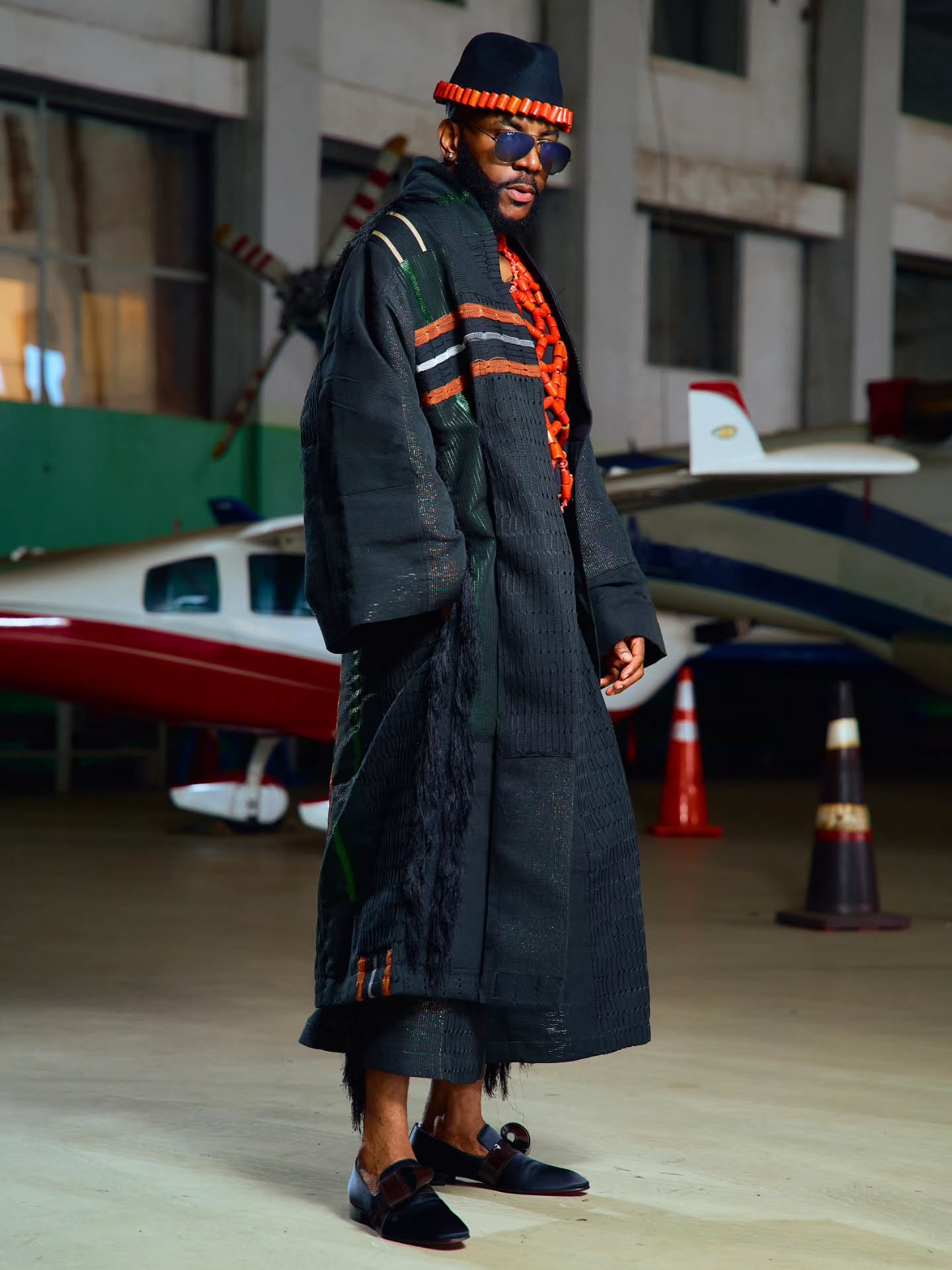 Ebuka Obi-Uchendu standing in an airfield hangar wearing a floor-length black Atafo coat, velvet loafers, and coral beads.
