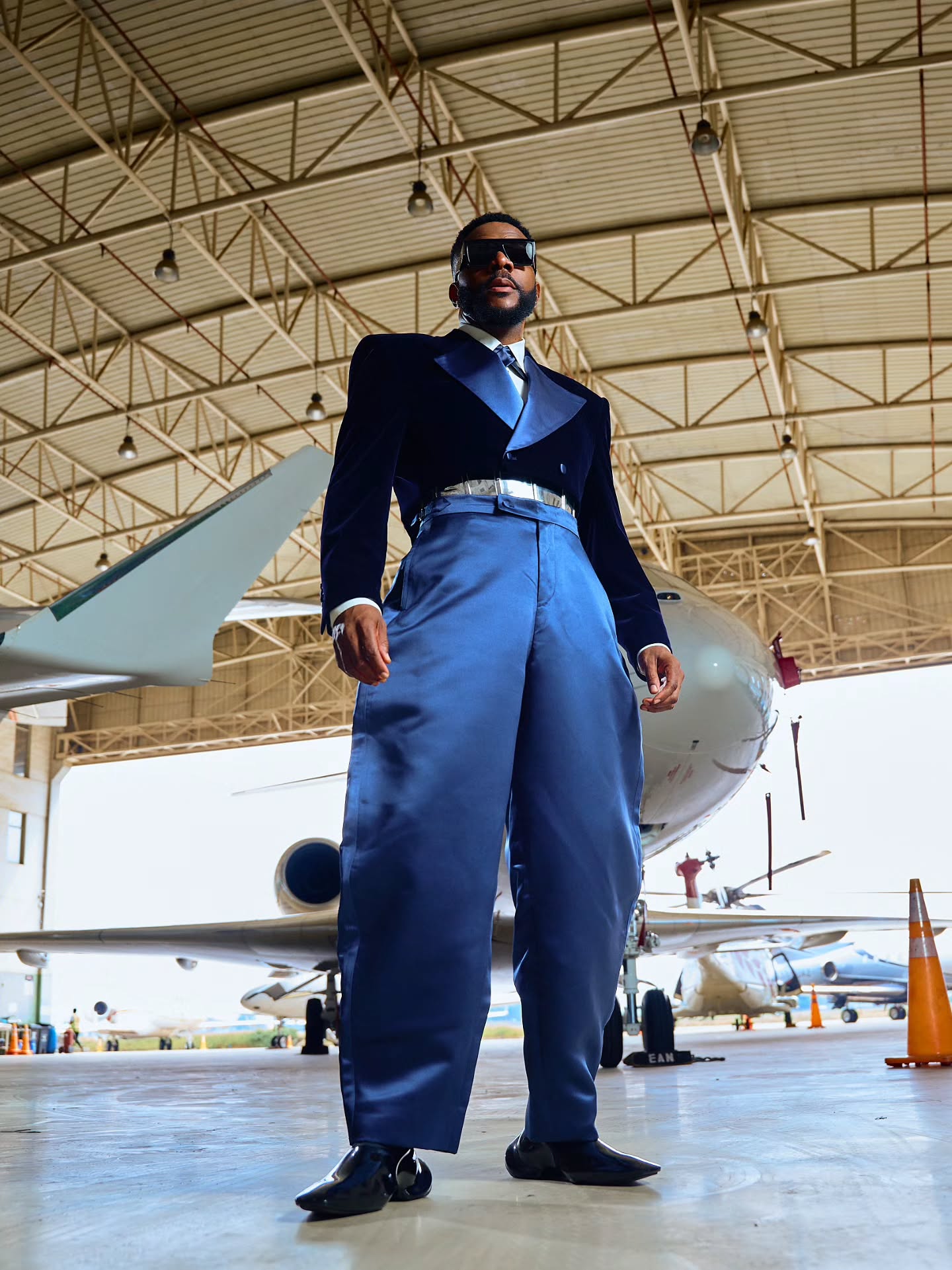 Ebuka Obi-Uchendu standing in an aircraft hangar in a Mai Atafo cropped velvet jacket and balloon trousers.