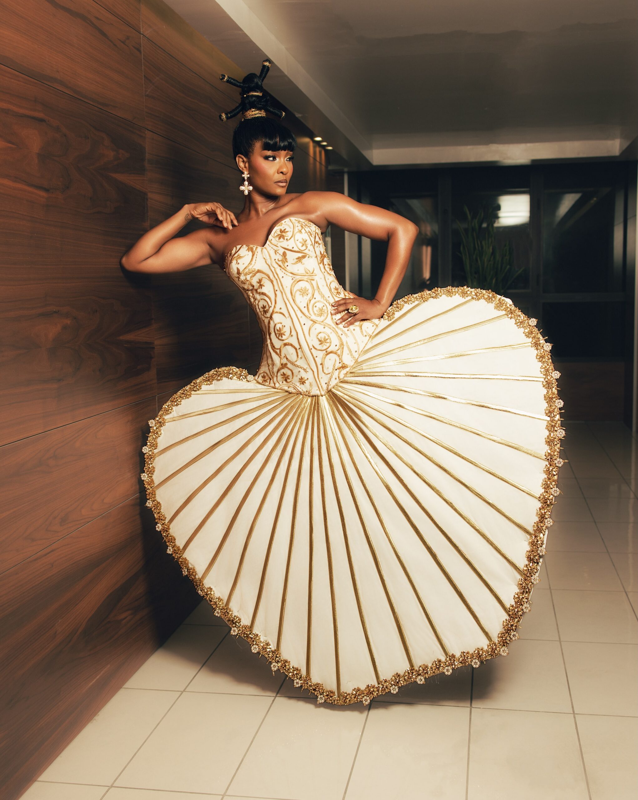 Osas Ighodaro leaning against a wooden wall wearing a heart-shaped fan skirt and gold-embroidered corset at BN20 Grand Red Carpet.