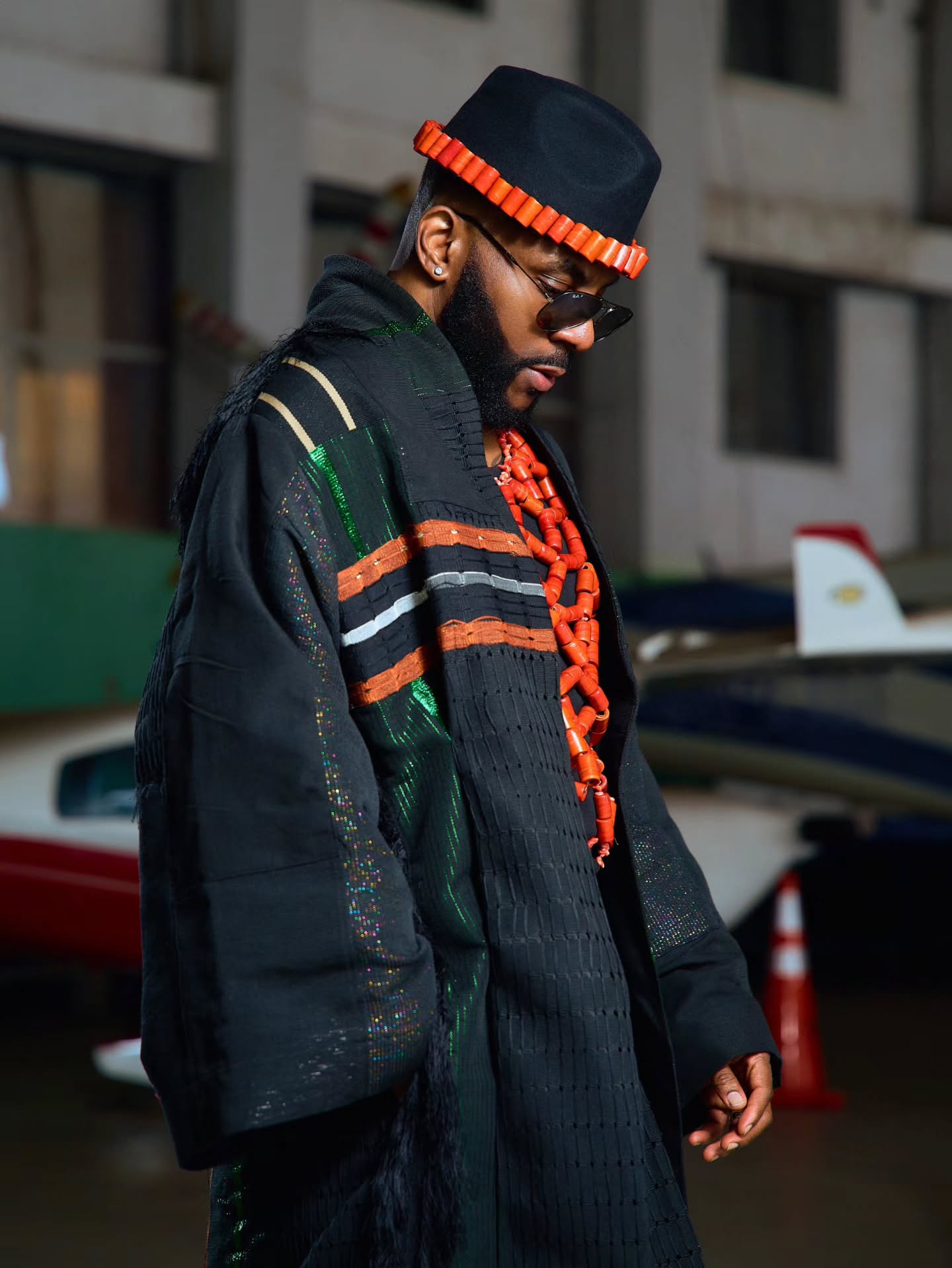 Ebuka Obi-Uchendu showing the coral-trimmed black fedora brim and multi-strand neck beads paired with an Atafo coat.