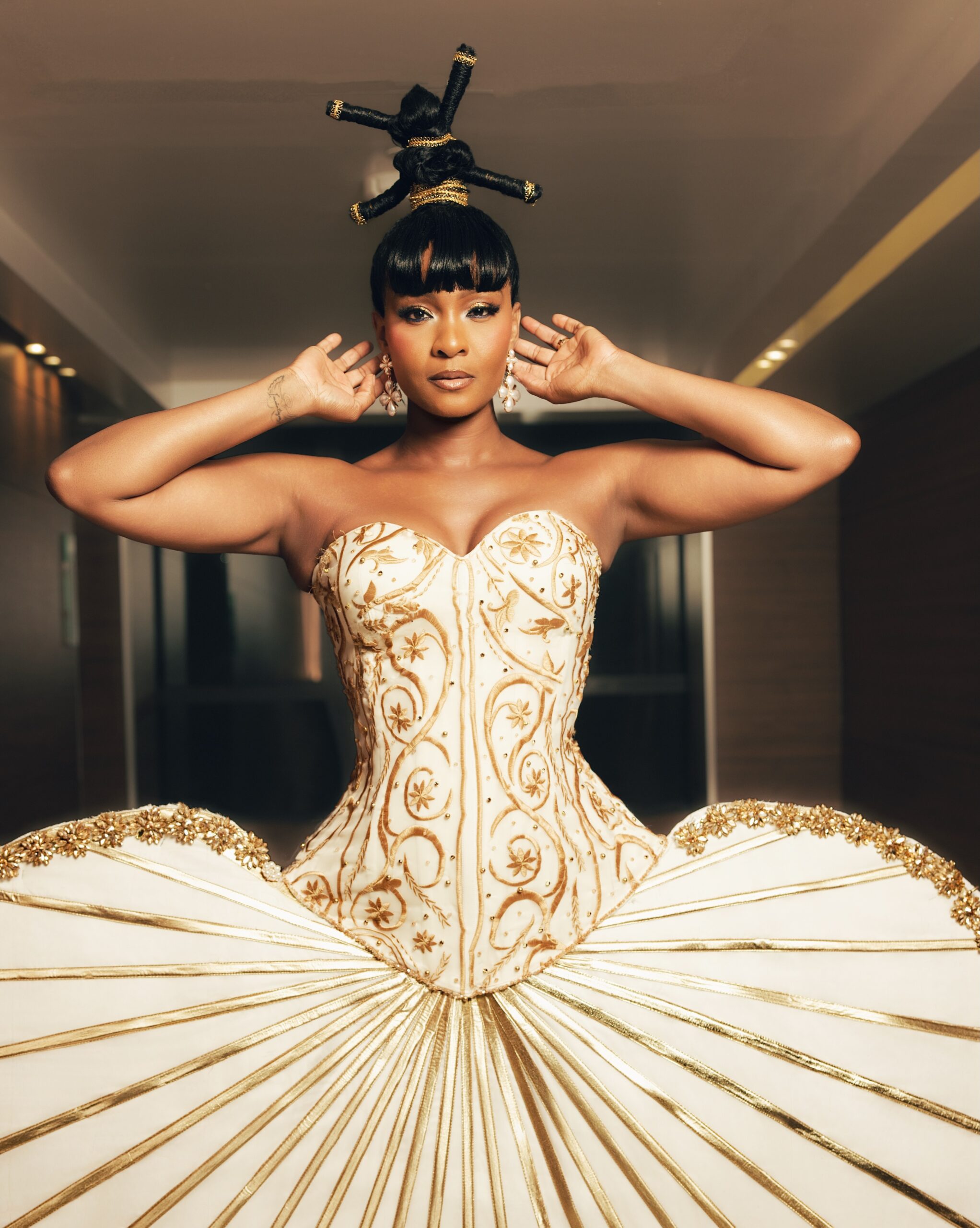 Osas Ighodaro posing in a custom ivory and gold corset dress with African threaded geometric knots for the BellaNaija at 20 Grand Red Carpet Experience.
