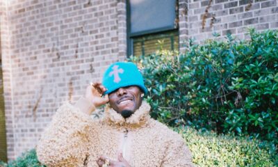 Asake wearing a beige fleece jacket, bright blue beanie, black leather trousers, and holding a round Louis Vuitton monogram hatbox bag.