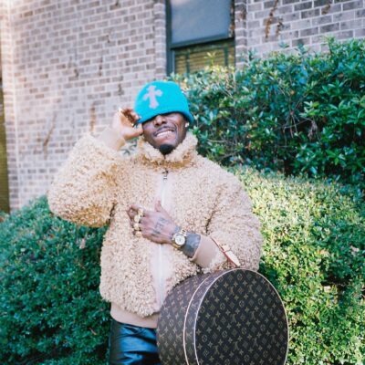 Asake wearing a beige fleece jacket, bright blue beanie, black leather trousers, and holding a round Louis Vuitton monogram hatbox bag.