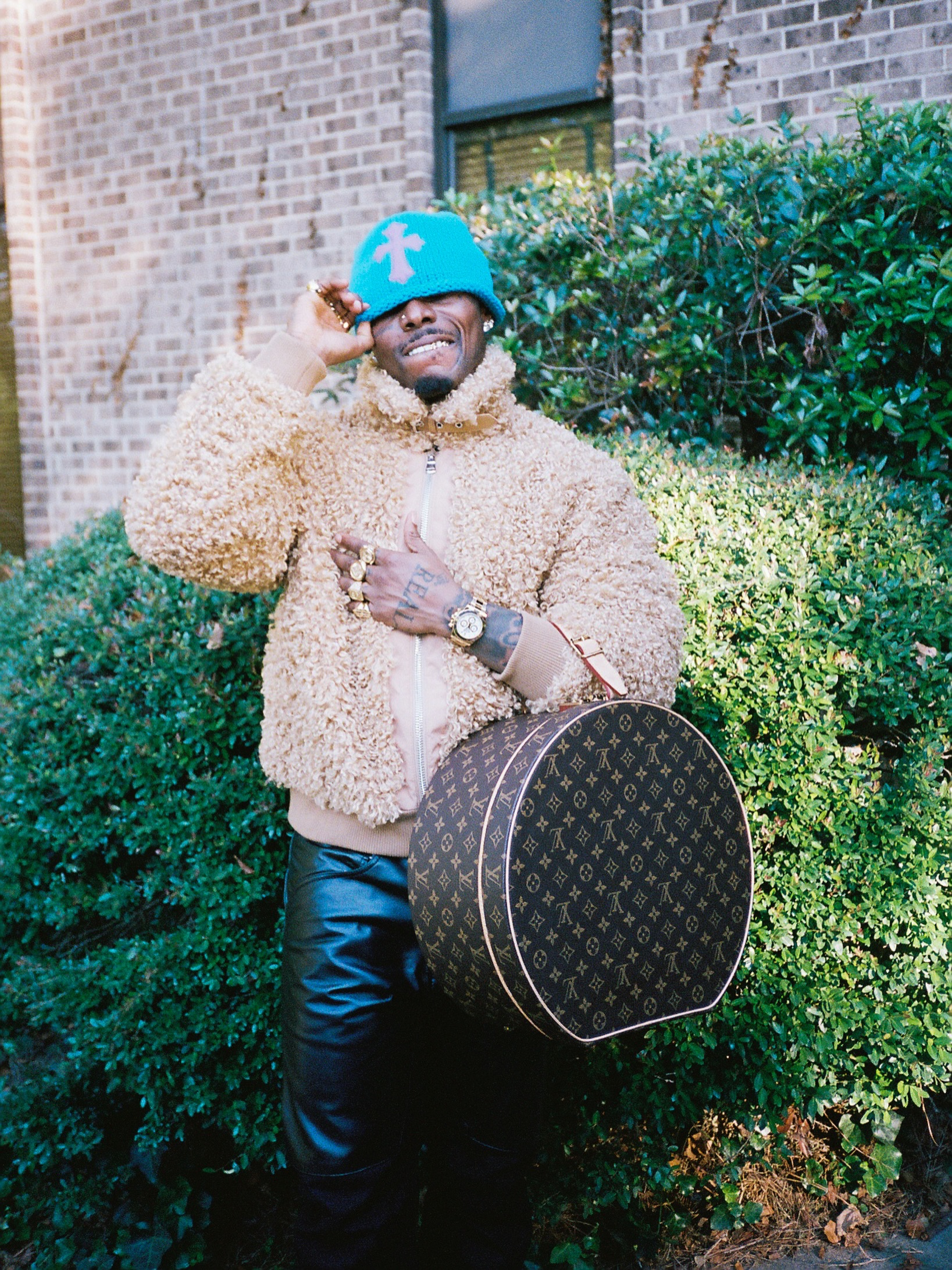 Asake wearing a beige fleece jacket, bright blue beanie, black leather trousers, and holding a round Louis Vuitton monogram hatbox bag.