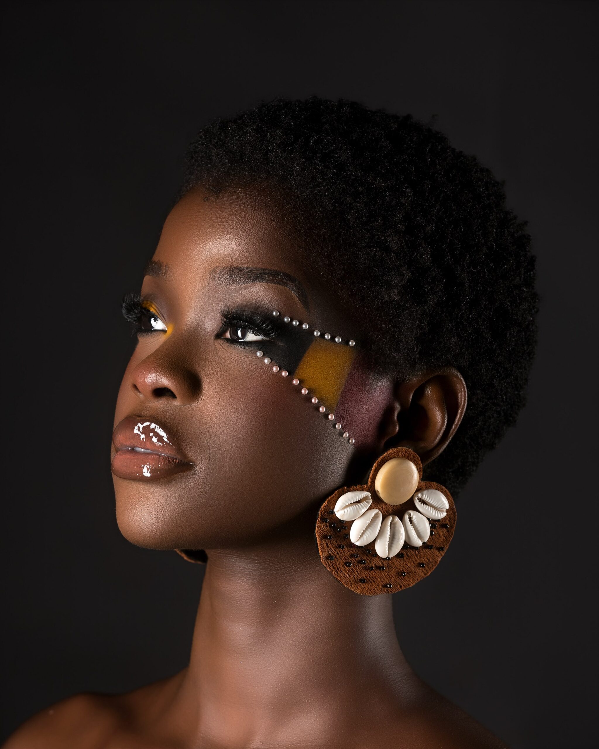 Kimberly Jael featuring graphic black, yellow, and red cheek makeup with pearl accents and cowrie shell drop earrings.