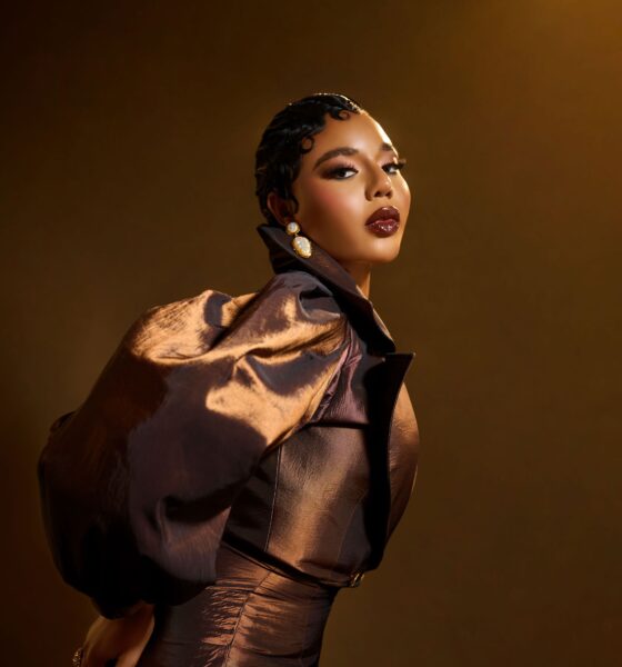 Nancy Isime featuring finger-wave hair, glossy dark lip liner, and pearl gold earrings in a metallic brown outfit.