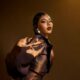 Nancy Isime featuring finger-wave hair, glossy dark lip liner, and pearl gold earrings in a metallic brown outfit.