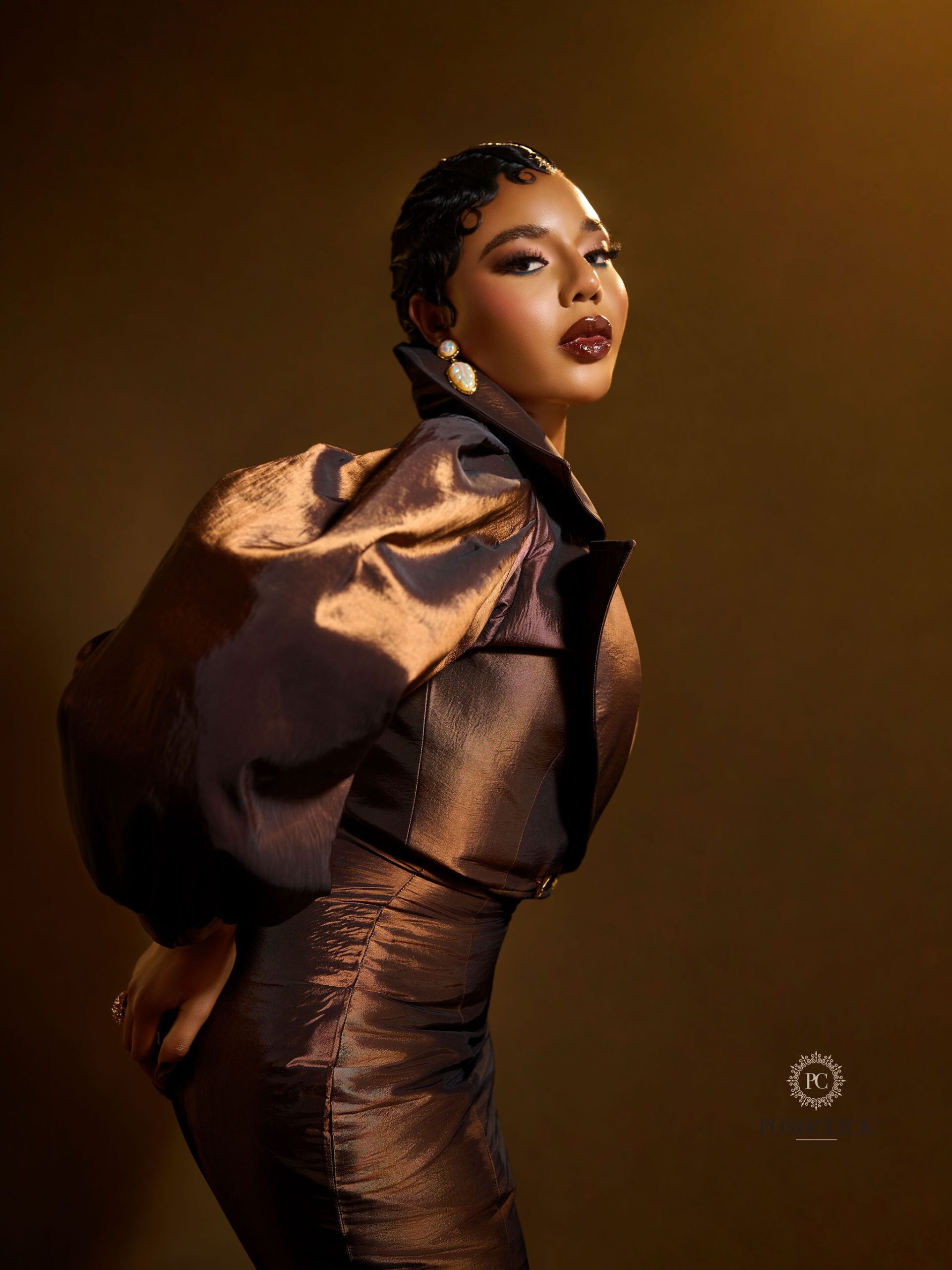 Nancy Isime featuring finger-wave hair, glossy dark lip liner, and pearl gold earrings in a metallic brown outfit.