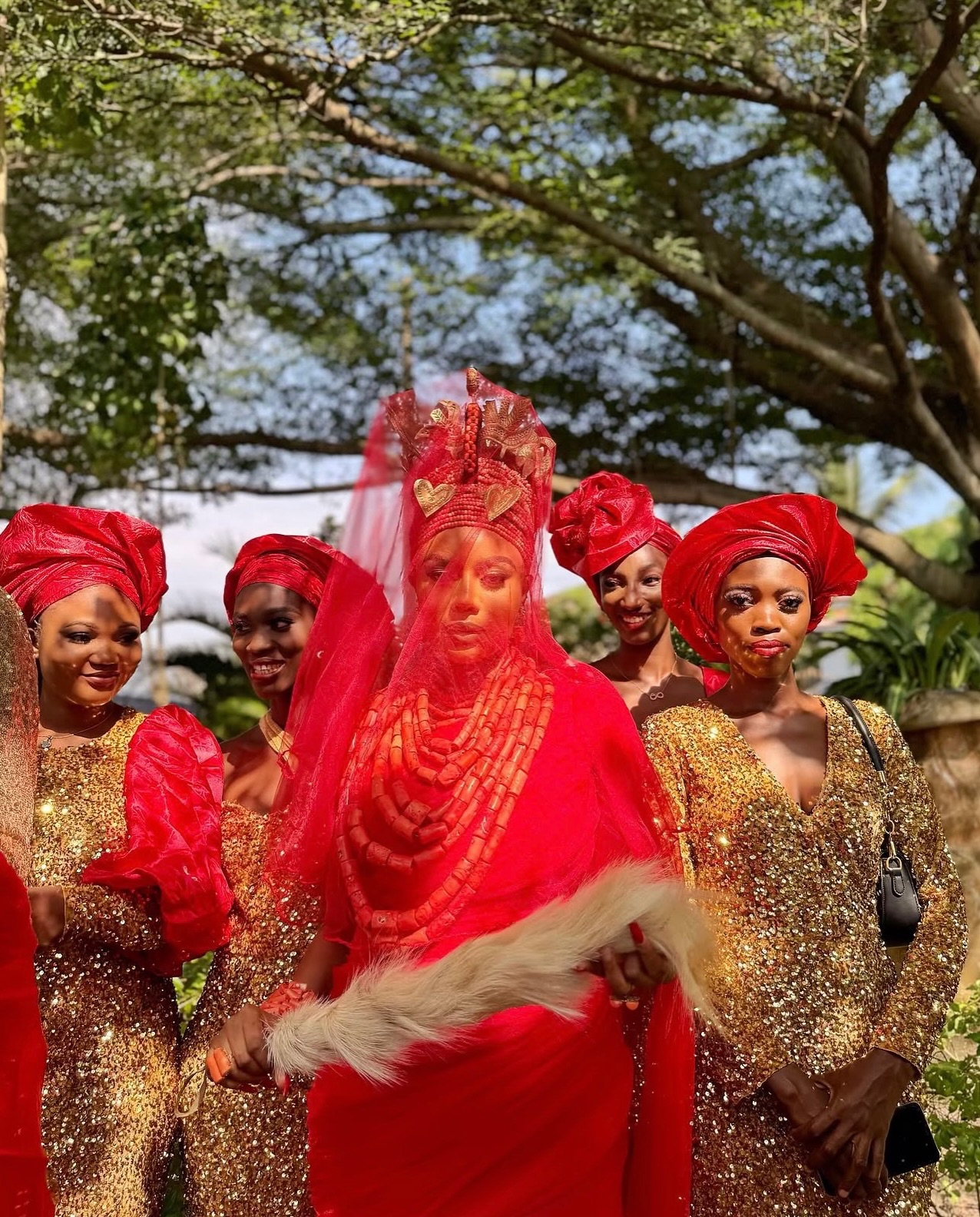Nancy Isime wearing a traditional red Edo outfit with Okuku coral bead crown and veil surrounded by women in sequined dresses on set of Loving Itohan.