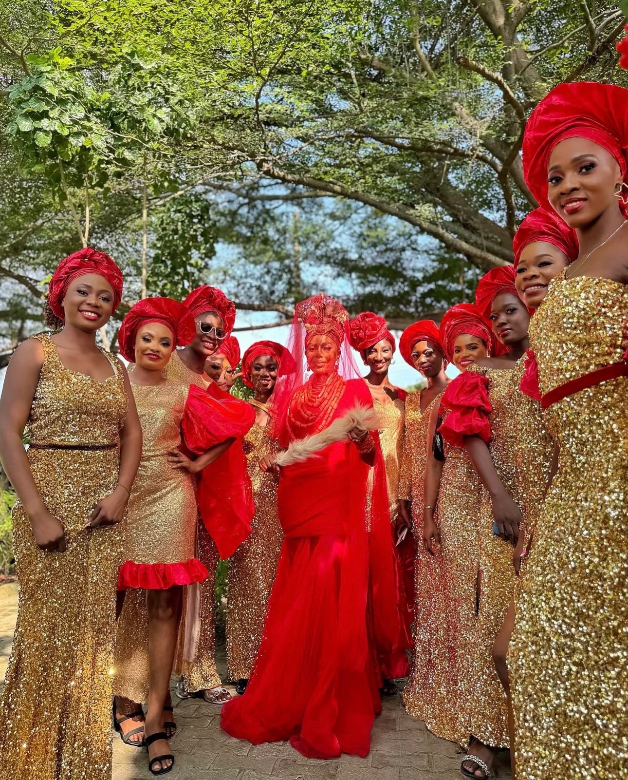 Nancy Isime in a red traditional outfit with a coral bead crown standing with women in gold sequined outfits outdoors.