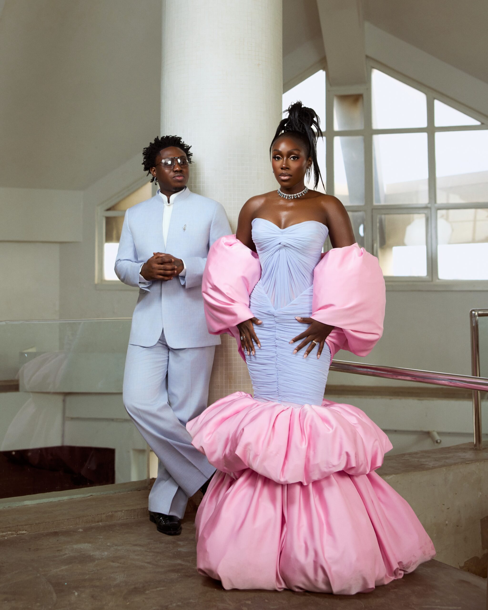 Zlatan Ibile in a light blue T.I Nathan suit leaning against a white pillar beside Davita Omoniyi in a Lasosa By Pam Pam gown with pink puff sleeves.