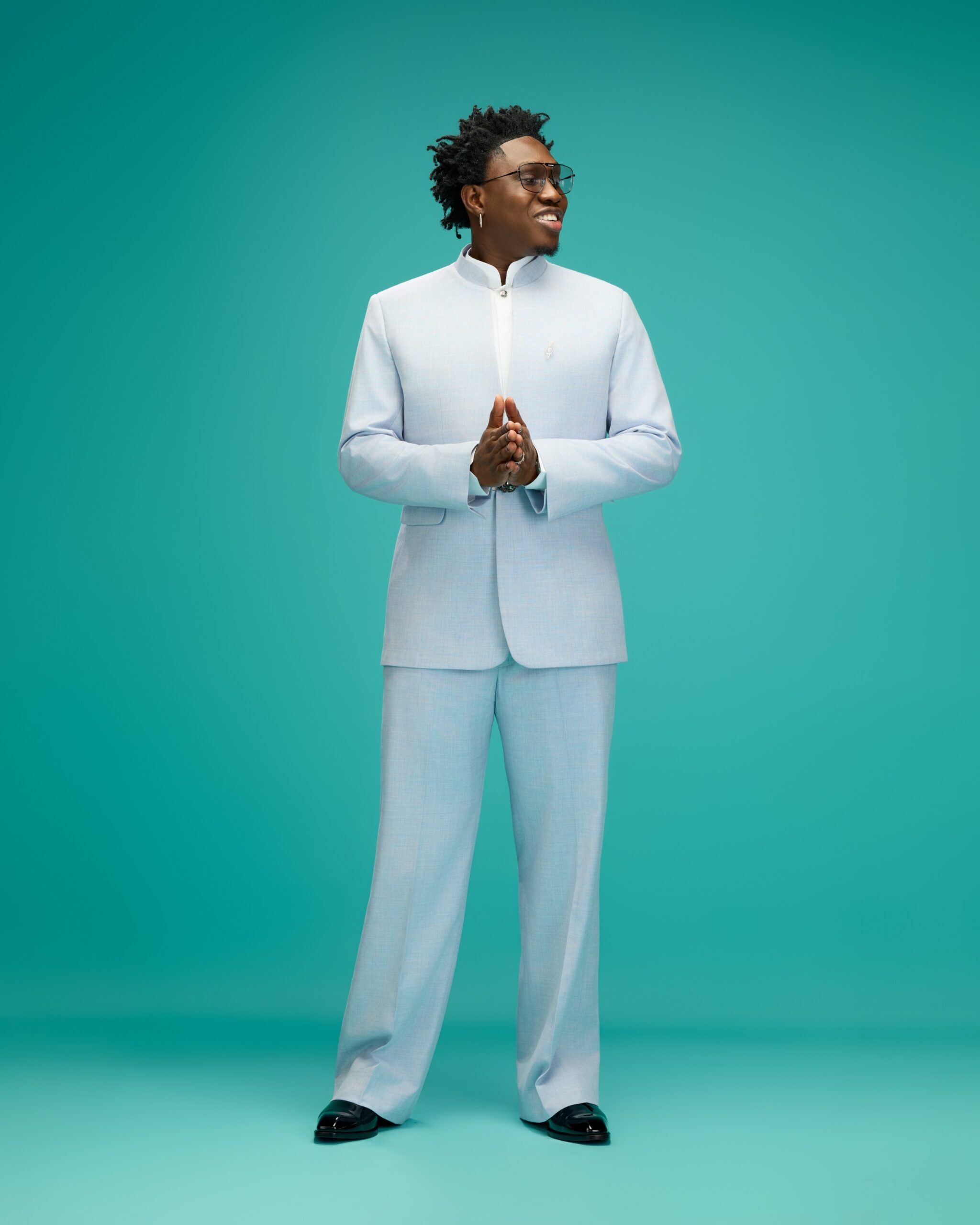 Zlatan Ibile standing with hands clasped wearing a light blue mandarin collar suit by T.I Nathan against a teal background.