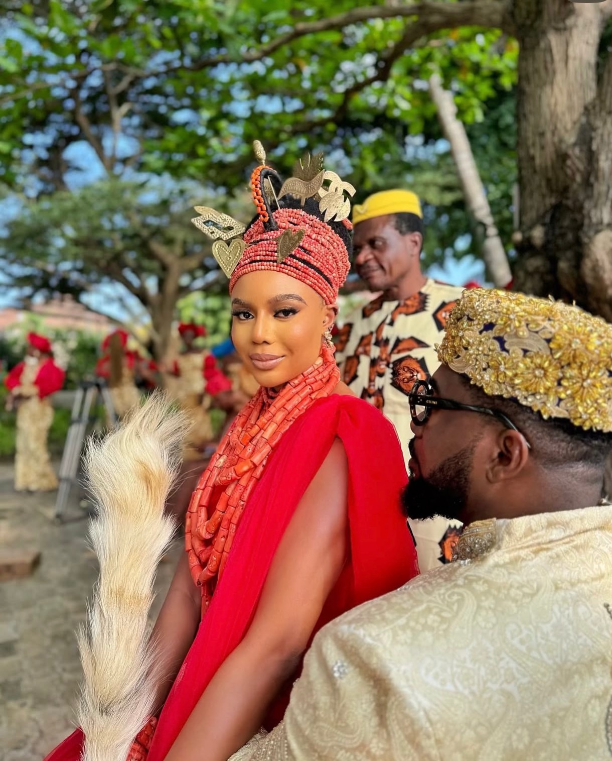 Nancy Isime wearing an Okuku coral bead crown looking at the camera while Uzor Arukwe looks towards her in a gold cap and glasses.