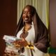 Saskay holding a stack of books wearing the off-white Hertunba Nola Kaftan with brown Aso-Oke appliques and a silk veil.