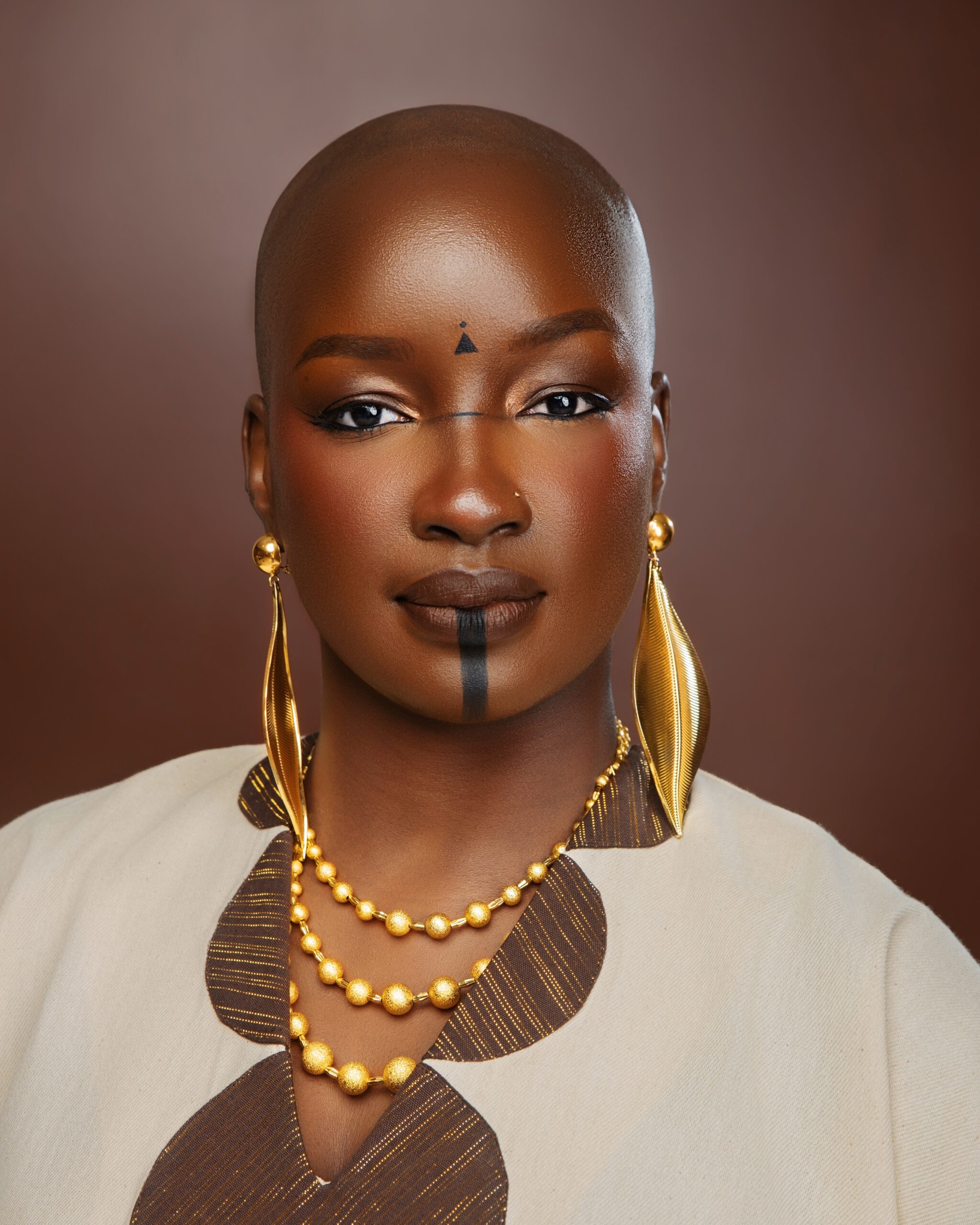 Saskay showing traditional African facial markings, oversized leaf-shaped gold drop earrings, and a layered beaded gold necklace.