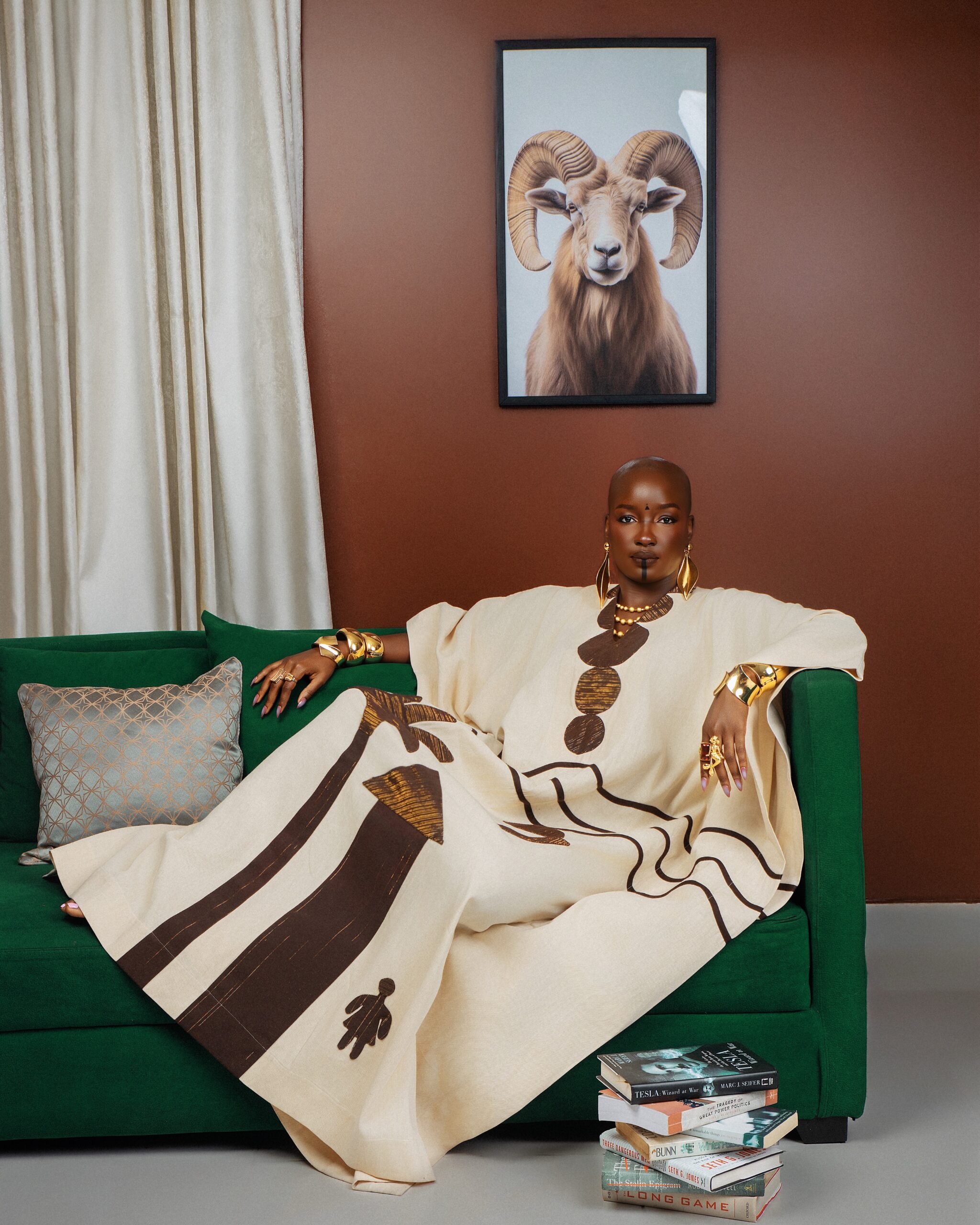 Saskay seated on a green couch in a flowing off-white kaftan with brown Aso-Oke appliques next to a stack of books.