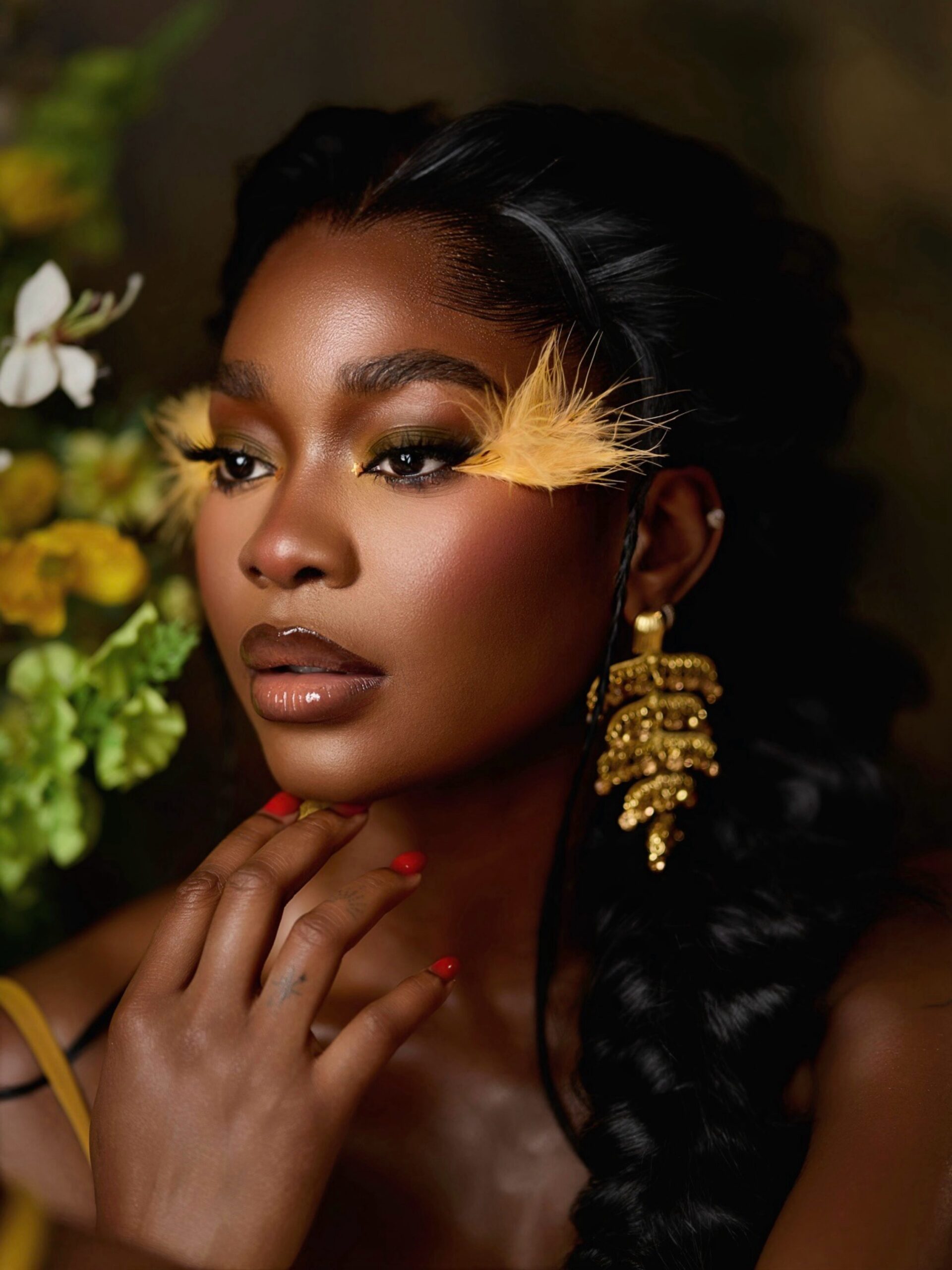 Dede Ashiogwu showing yellow feather-accented eye makeup, glossy nude lips, and gold leaf drop earrings.