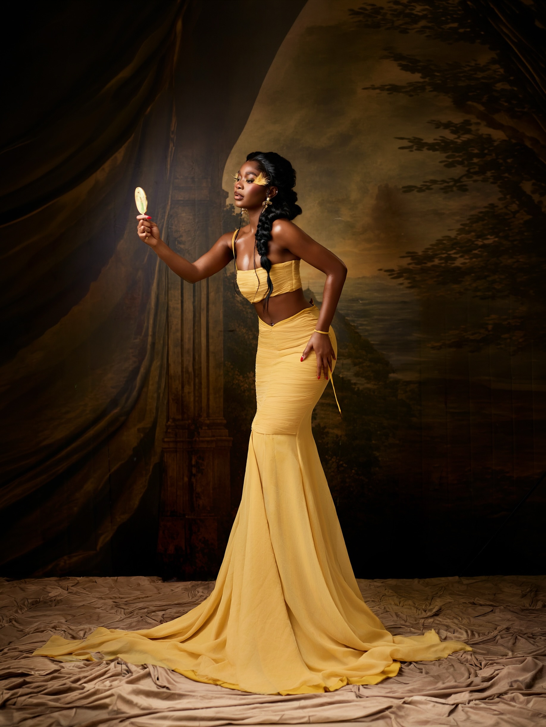 Dede Ashiogwu holding a vintage gold handheld mirror in a butter yellow two-piece set.