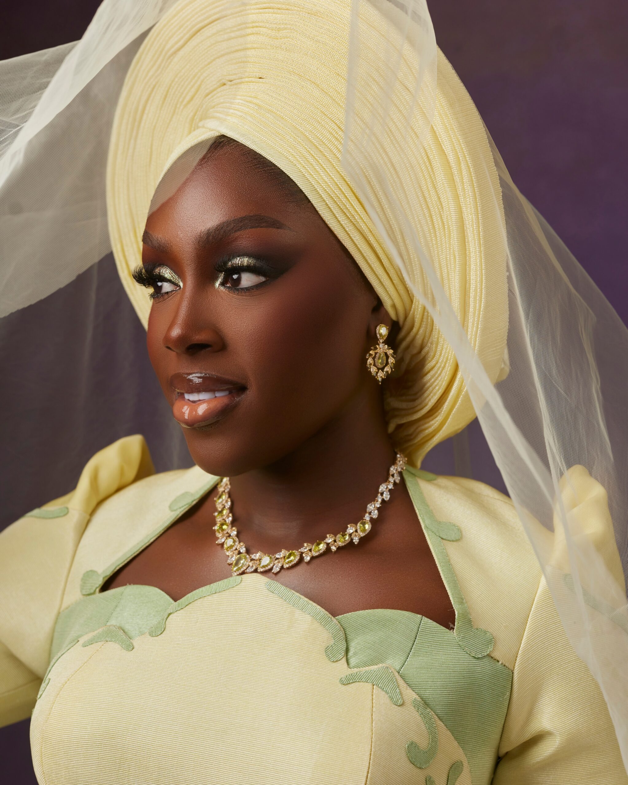 Davita Omoniyi wearing a pale lemon-yellow pleated gele headwrap with a sheer tulle veil, olive-green gemstone necklace, and bridal makeup.