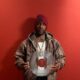 Skepta wearing a camouflage zip-up jacket and Supreme beanie holding a clear vinyl copy of his album The Fork and Knife against a red background.