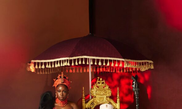 Zlatan Ibile holding a wooden staff seated on a gold throne beneath a tasseled umbrella next to Davita Omoniyi in Edo bridal attire.