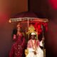 Zlatan Ibile holding a wooden staff seated on a gold throne beneath a tasseled umbrella next to Davita Omoniyi in Edo bridal attire.
