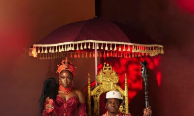 Zlatan Ibile holding a wooden staff seated on a gold throne beneath a tasseled umbrella next to Davita Omoniyi in Edo bridal attire.