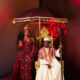 Zlatan Ibile holding a wooden staff seated on a gold throne beneath a tasseled umbrella next to Davita Omoniyi in Edo bridal attire.