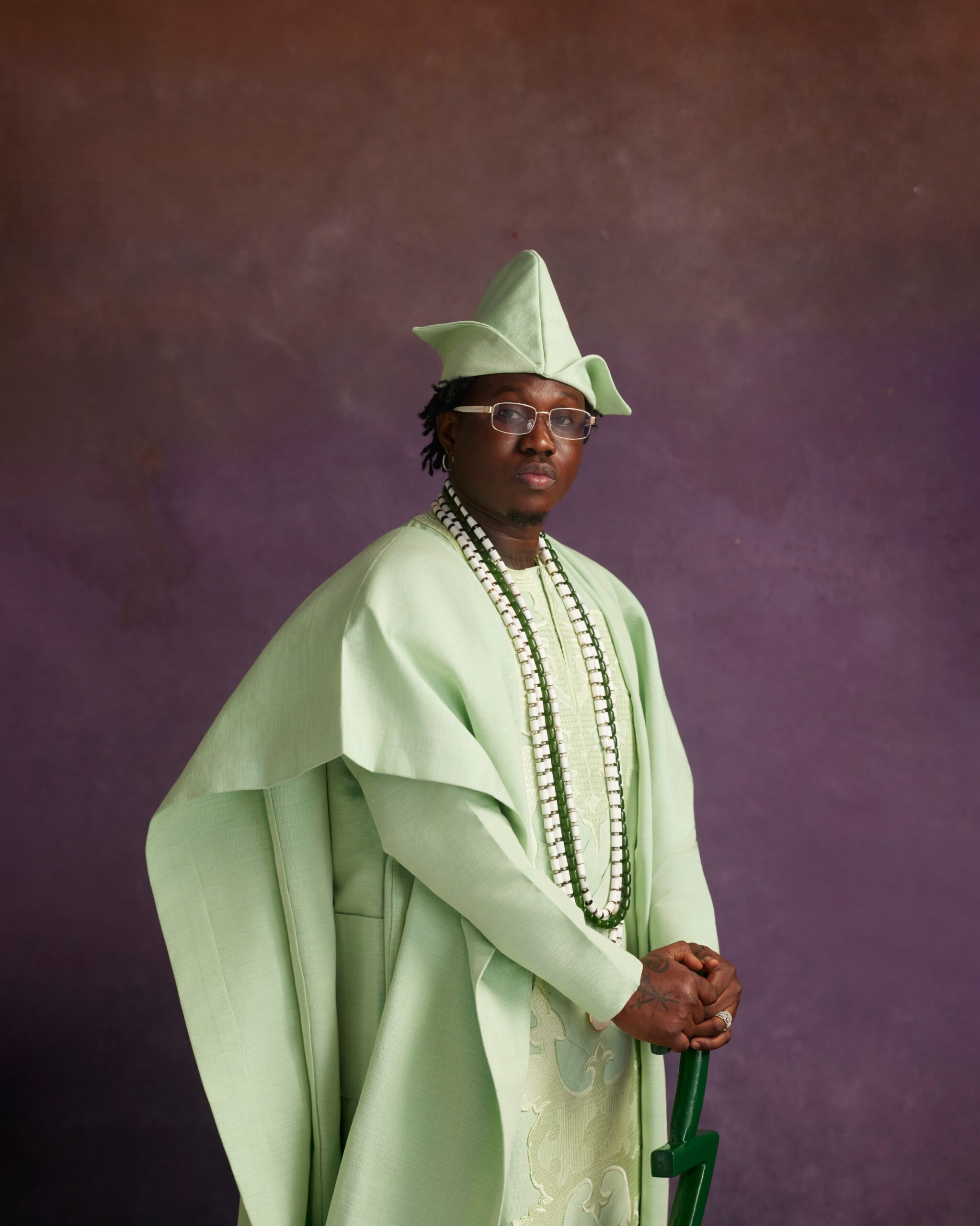 Zlatan Ibile holding a carved dark green walking stick wearing a mint-green Agbada, matching Fila cap, and layered white beaded necklaces.