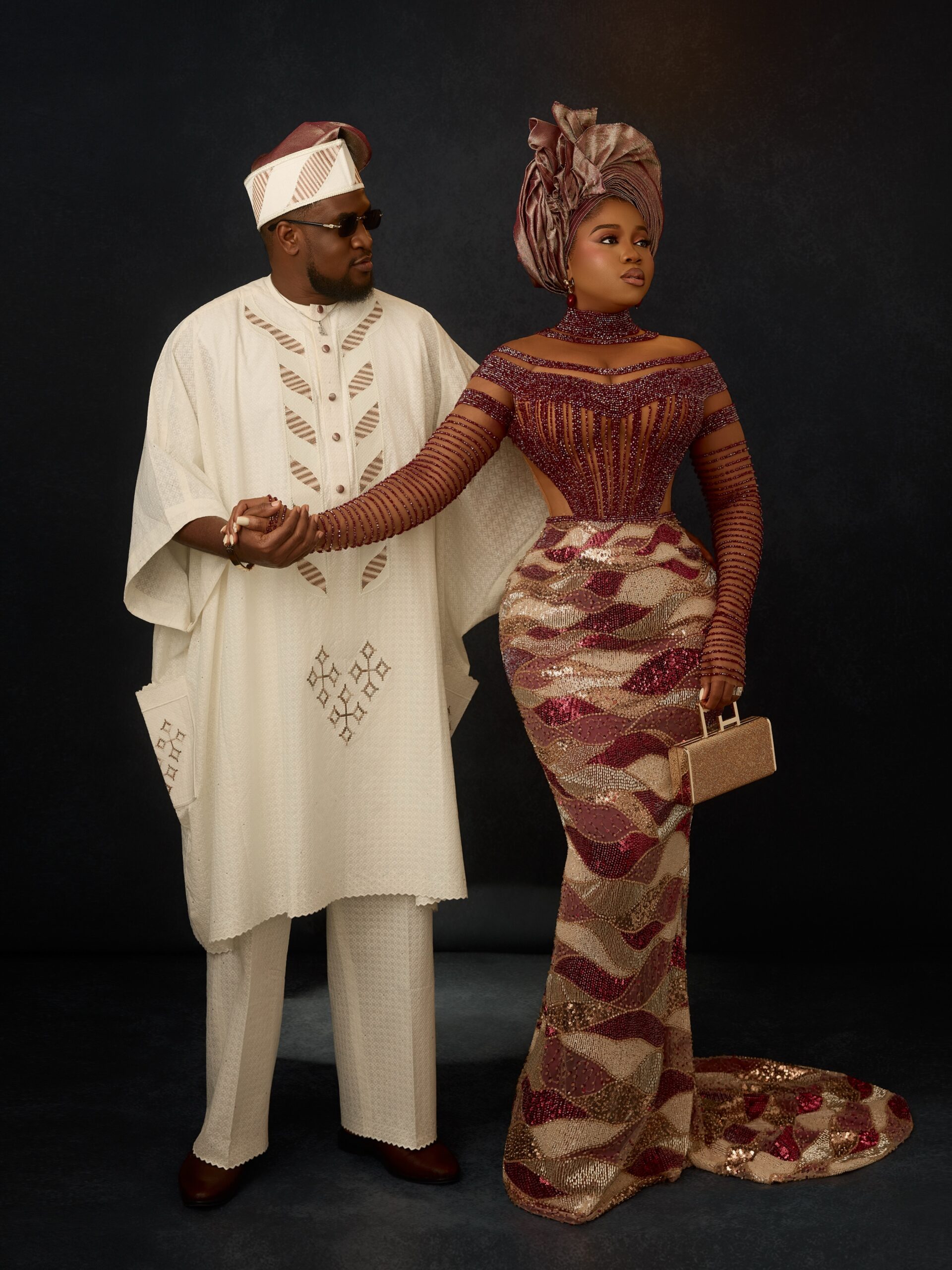 Veekee James in a wine-red sheer corset dress holding hands with her husband Femi Atere in an off-white Agbada and matching Fila cap.