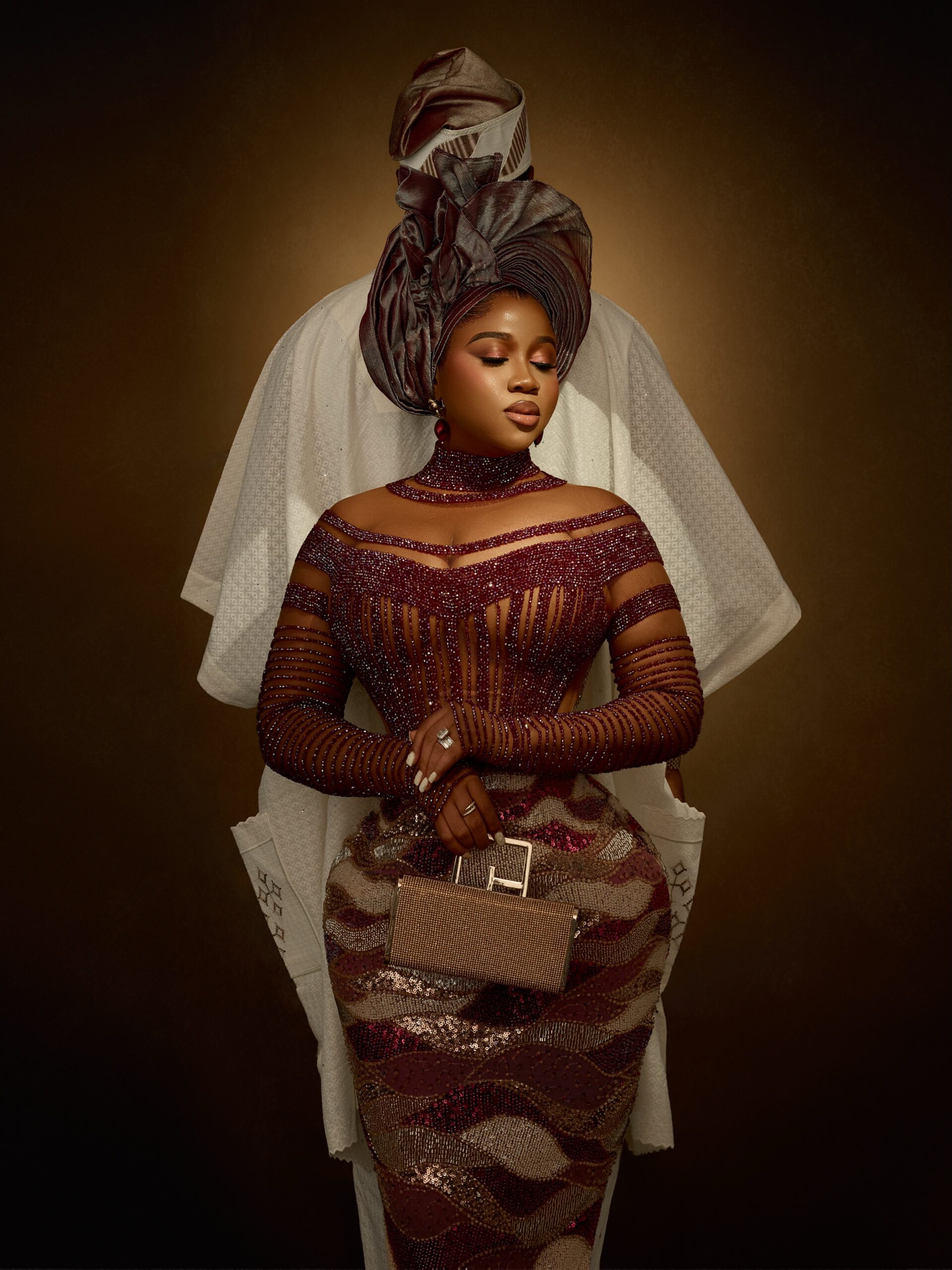 Veekee James posing in a wine-red corset gown with her husband Femi Atere standing behind her in an off-white Agbada.