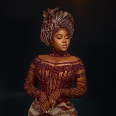 Veekee James wearing a high-neck wine red crystal choker, sheer illusion corset bodice, and pleated metallic dusty rose gele.