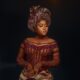 Veekee James wearing a high-neck wine red crystal choker, sheer illusion corset bodice, and pleated metallic dusty rose gele.