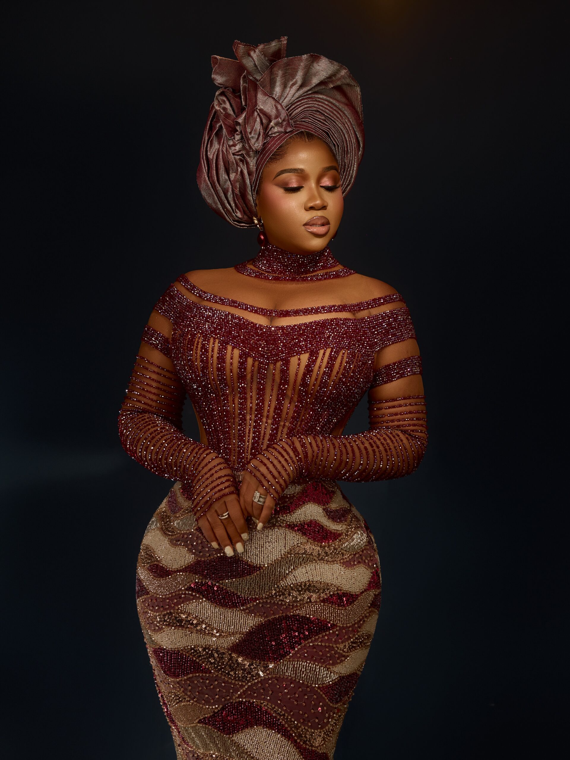 Veekee James wearing a high-neck wine red crystal choker, sheer illusion corset bodice, and pleated metallic dusty rose gele.